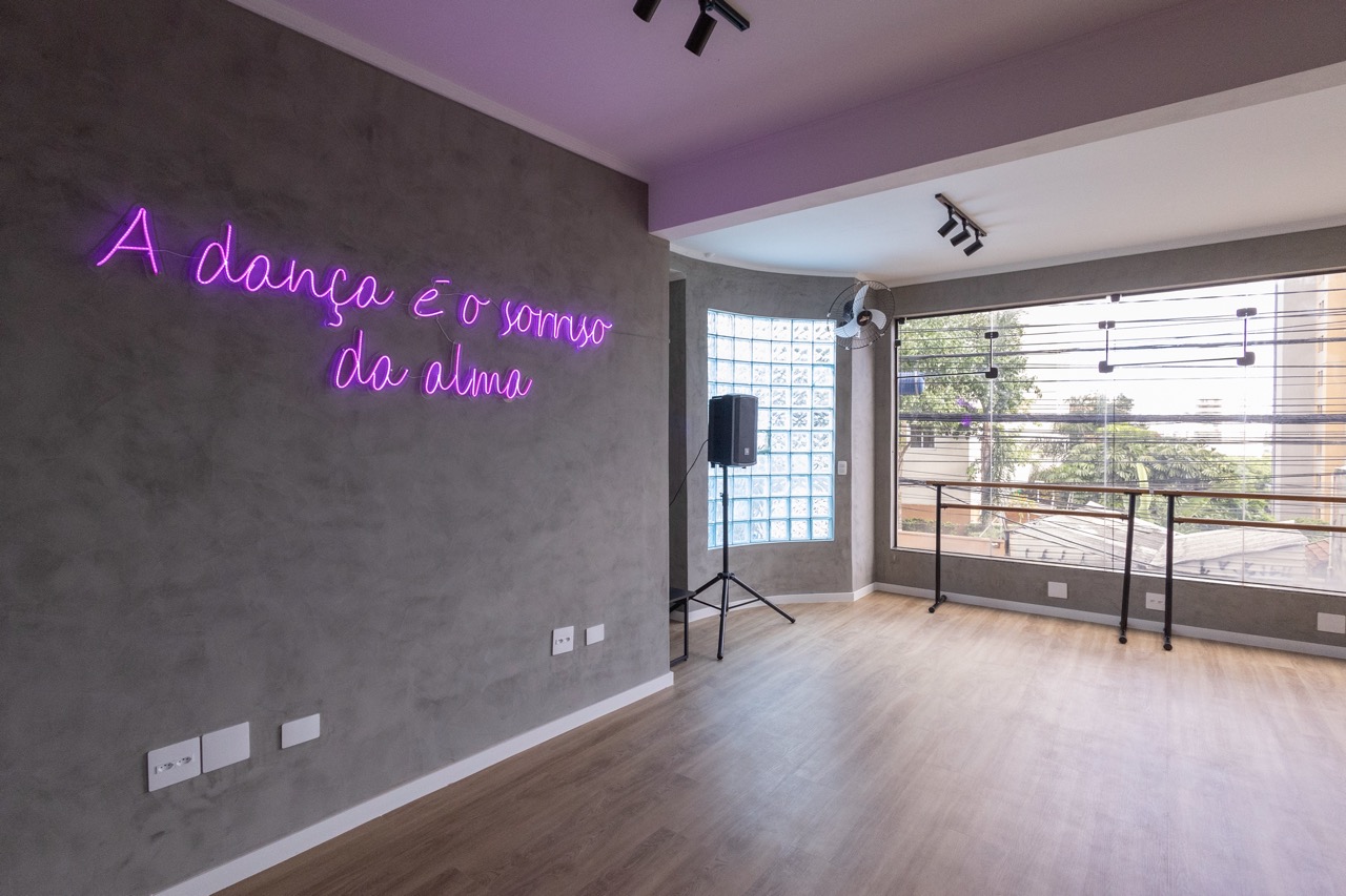 Sala de dança do Estúdio Dancefloor com piso de madeira, barra de ballet e neon 'A dança é o sorriso da alma'